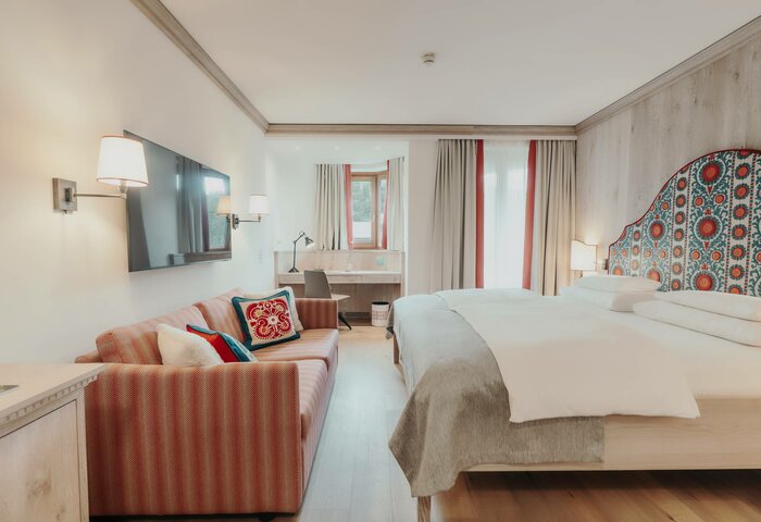 Zimmer in einem der schönsten Hotels in Kärnten am See Ein helles Hotelzimmer mit einem großen Doppelbett, einem gemütlichen Sofa in Rot- und Beigetönen, einem Schreibtisch und einem Fernseher an der Wand. Große Fenster mit Vorhängen lassen viel Tageslicht herein. Die Wände sind in warmen Holztönen gehalten.