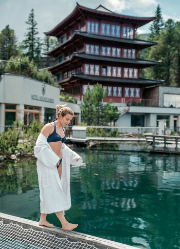 Urlaubsangebote für das ganze Jahr - Turracher Höhe Eine Frau in einem weißen Bademantel und Bikini steht am Rand eines klaren Gewässers. Im Hintergrund ist ein mehrstöckiges, traditionell gestaltetes Gebäude mit roten Akzenten und umgebenden Bäumen zu sehen. Der Himmel ist teilweise bewölkt.