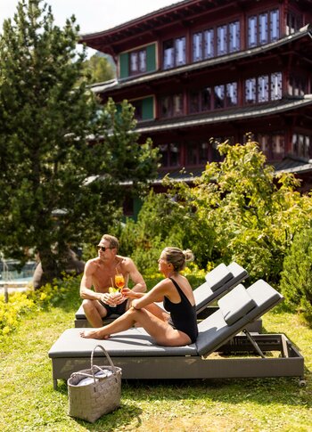 Urlaubsangebote für das ganze Jahr - Turracher Höhe Ein Paar sitzt entspannt auf Liegen im Freien, umgeben von grünen Pflanzen und Bäumen. Sie genießen Cocktails und unterhalten sich. Im Hintergrund ist ein großes, traditionelles Gebäude mit mehreren Etagen sichtbar.