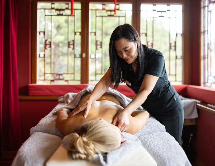 Chinese head and back massage at Kristall Spa A therapist massages the back of a reclining woman in a treatment room with red curtains and large windows.