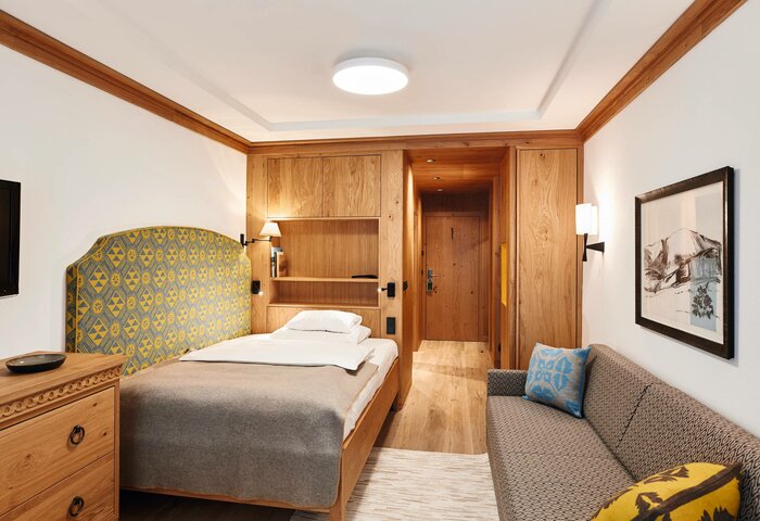 Alles für Genuss-Urlaub auf der Turracher Höhe, Kärnten. Ein modernes Hotelzimmer mit einem Einzelbett, einem gepolsterten Kopfteil in Gelb und Blau, einem Holztisch und einer Couch. Die Wände sind hell, und es gibt Holzverkleidungen. Ein Gemälde hängt an der Wand, und die Tür zum Flur ist sichtbar.