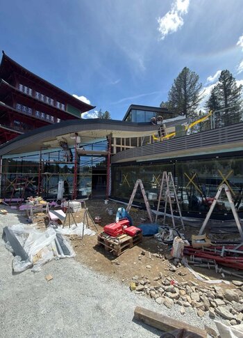 Mitten in Kärnten Hotel am See. 4-Sterne-Superior-Genuss Bauarbeiten an einem modernen Gebäude mit großen Fenstern und einer geschwungenen Überdachung. Im Vordergrund sind Baustellenmaterialien, Gerüste und unebener Boden sichtbar. Im Hintergrund steht ein traditionelles Gebäude mit rotem Dach und Bäumen. Der Himmel ist blau mit einigen Wolken.