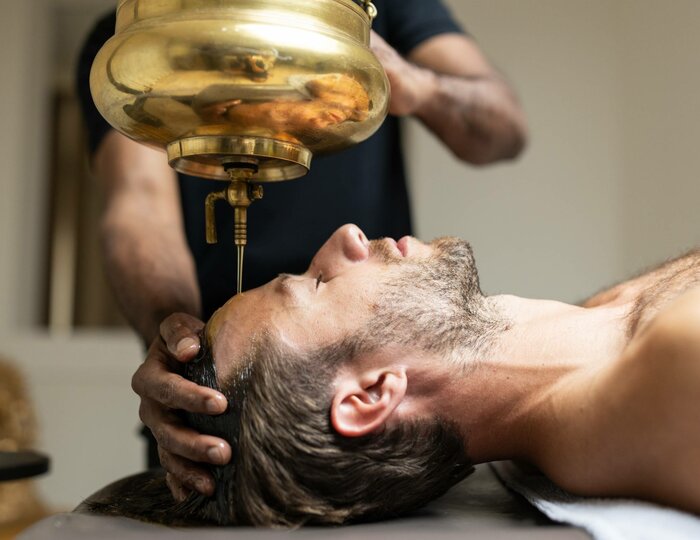 Shirodhara treatment at Kristall Spa A therapist supports a man's head while oil flows from a brass vessel onto his forehead.