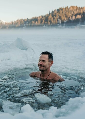 Urlaubsangebote für das ganze Jahr - Turracher Höhe Ein Mann steht in einem offenen Wasserloch, umgeben von Schnee und Eis. Er lächelt und genießt die kalte Umgebung. Im Hintergrund sind schneebedeckte Berge und Bäume zu sehen, während der Himmel klar ist.
