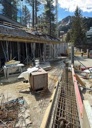 Mitten in Kärnten Hotel am See. 4-Sterne-Superior-Genuss Baugelände mit einem Arbeiter, der in der Nähe von Baustellenmaterial und Stahlträgern steht. Im Hintergrund sind Bäume und ein Gebäude sichtbar. Der Himmel ist teilweise bewölkt.