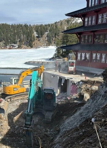 Mitten in Kärnten Hotel am See. 4-Sterne-Superior-Genuss Bauarbeiten am Ufer eines gefrorenen Sees, mit zwei Baggern, die Erde und Steine bewegen. Im Hintergrund ist ein mehrstöckiges Gebäude mit roten Fenstern und eine bewaldete Hügelkette sichtbar. Der Himmel ist teilweise bewölkt.