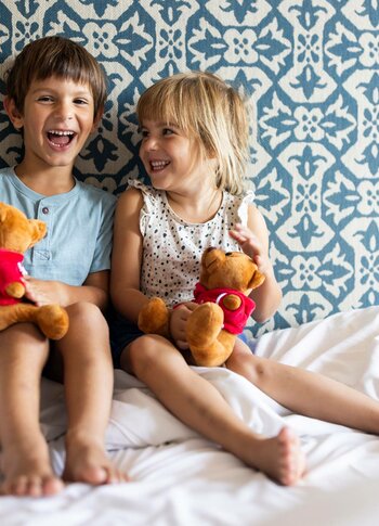 Kinder auf dem Hotelbett im Familienzimmer Zwei Kinder sitzen lachend auf dem Bett in einem hellen Hotelzimmer und halten Plüschtiere in den Händen.