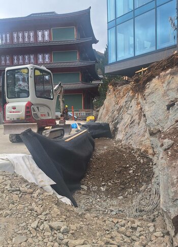 Mitten in Kärnten Hotel am See. 4-Sterne-Superior-Genuss Blick auf eine Baustelle mit einem Bagger, der an einem Felsen arbeitet. Im Hintergrund ist ein modernes Gebäude mit großen Fenstern und ein traditionelles Gebäude mit Holzverzierungen zu sehen. Der Boden ist mit Steinen und schwarzem Material bedeckt.