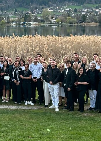 Mitten in Kärnten Hotel am See. 4-Sterne-Superior-Genuss Eine große Gruppe von Menschen steht auf einer Wiese am Ufer eines Sees, umgeben von hohen Schilfpflanzen. Die Teilnehmer tragen überwiegend schwarze und weiße Kleidung. Im Hintergrund sind sanfte Hügel und eine kleine Siedlung sichtbar.