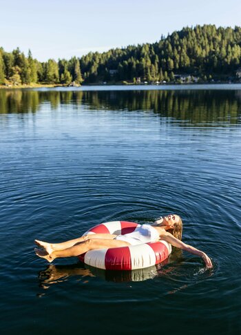 Urlaubsangebote für das ganze Jahr - Turracher Höhe Eine Person liegt entspannt auf einem roten und weißen Schwimmreifen im ruhigen Wasser eines Sees. Umgeben von grünen Bäumen und sanften Hügeln, reflektiert das Wasser die klare blaue Himmel. Die Atmosphäre ist friedlich und einladend.