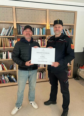 Mitten in Kärnten Hotel am See. 4-Sterne-Superior-Genuss Zwei Männer stehen in einem Raum mit Bücherregalen im Hintergrund. Der Mann links trägt eine schwarze Jacke und Jeans, während der Mann rechts in einer Feuerwehruniform mit Mütze steht. Beide halten ein großes Schild mit der Aufschrift „SPENDENCHECK 4.304“.