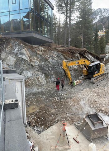 Mitten in Kärnten Hotel am See. 4-Sterne-Superior-Genuss Blick auf eine Baustelle mit einem Bagger, der an einem steilen, felsigen Hang arbeitet. Zwei Arbeiter stehen in der Nähe des Baggers. Im Hintergrund sind Bäume und schneebedeckte Berge sichtbar. Ein modernes Gebäude mit großen Fenstern ist ebenfalls im Bild.